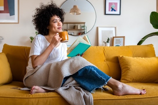 Une personne assise sur un divan tient une tasse et un livre.